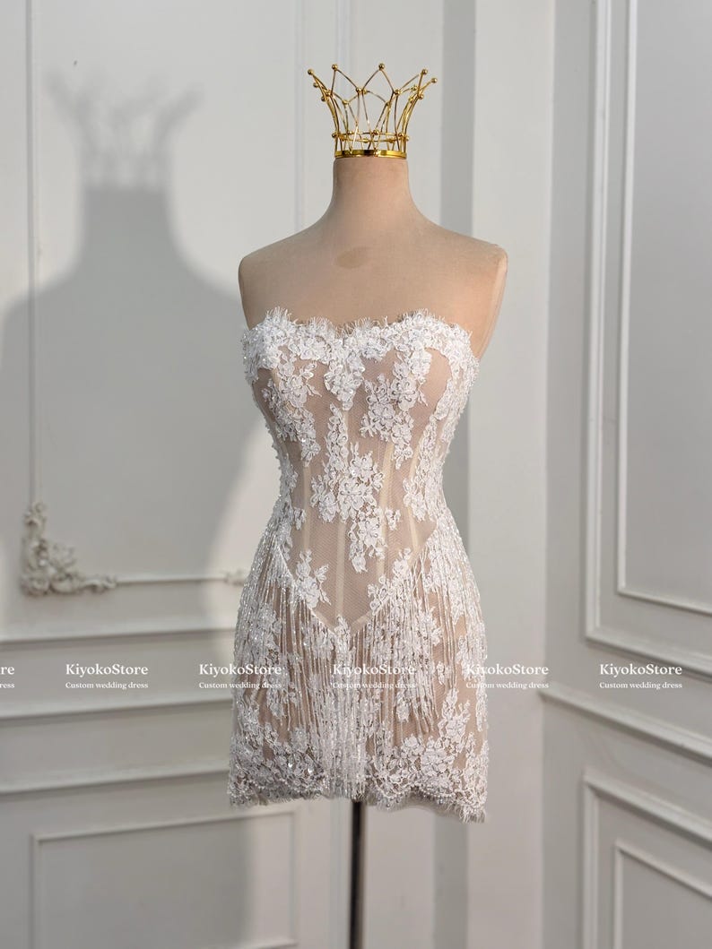 May include: A strapless mini dress featuring a nude-colored base with white floral lace overlays, intricate embroidery, and a fringe hemline. A gold-toned wire crown sits atop the mannequin displaying the dress, which includes the text KiyokoStore.