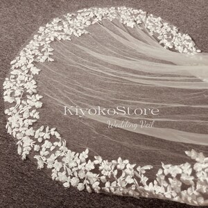 May include: A white wedding veil with delicate floral lace trim. The veil is made of soft tulle and has a scalloped edge. The text "KiyokoStore Wedding Veil" is printed on the veil.