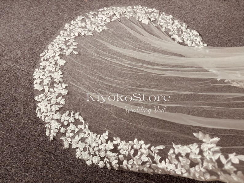 May include: A white wedding veil with delicate floral lace trim. The veil is made of soft tulle and has a scalloped edge. The text "KiyokoStore Wedding Veil" is printed on the veil.