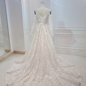 May include: A white off-the-shoulder wedding dress with long sleeves and a star-shaped pattern. The dress has a fitted bodice and a flowing skirt.