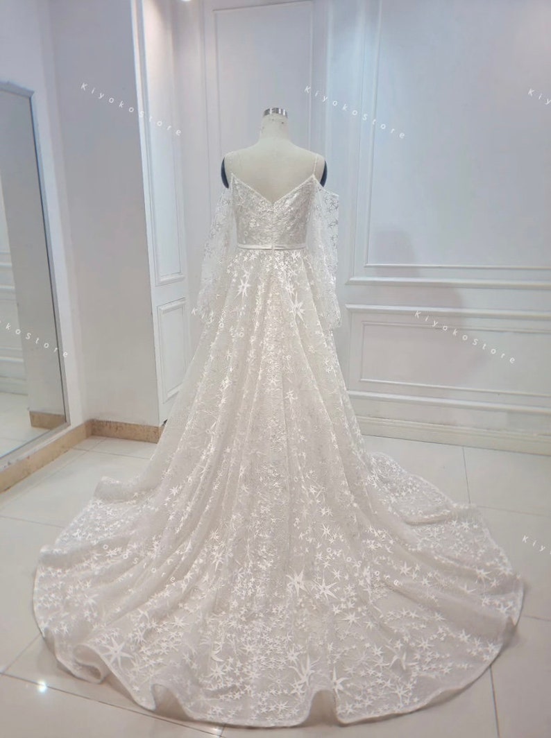 May include: A white off-the-shoulder wedding dress with long sleeves and a star-shaped pattern. The dress has a fitted bodice and a flowing skirt.