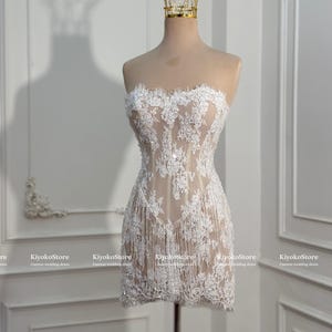 May include: A strapless mini dress featuring white floral lace embroidery over a nude-colored base. The garment includes a fringe hemline and subtle sequin embellishments throughout the lace pattern. A gold-colored crown sits atop the mannequin.