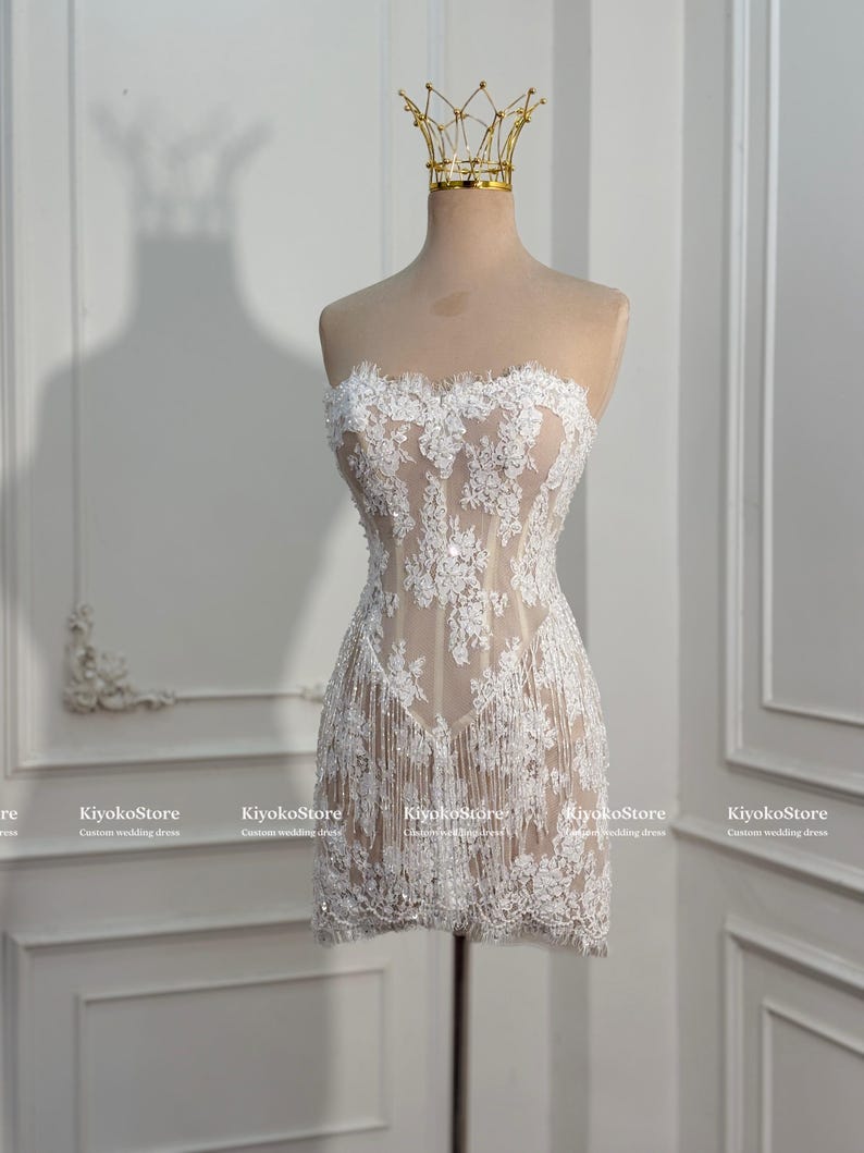 May include: A strapless mini dress featuring white floral lace embroidery over a nude-colored base. The garment includes a fringe hemline and subtle sequin embellishments throughout the lace pattern. A gold-colored crown sits atop the mannequin.