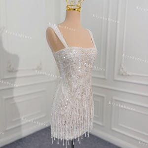 May include: A white, beaded mini dress with a square neckline and spaghetti straps. The dress is adorned with sequins and dangling fringe. A gold crown sits atop the mannequin's head. The dress is displayed on a wooden stand.