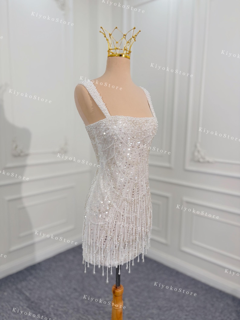 May include: A white, beaded mini dress with a square neckline and spaghetti straps. The dress is adorned with sequins and dangling fringe. A gold crown sits atop the mannequin's head. The dress is displayed on a wooden stand.