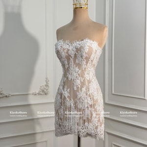 May include: A strapless mini dress featuring a nude-colored base with white floral lace overlays and delicate fringe detailing along the hem. The garment is displayed on a mannequin wearing a gold-colored crown-shaped headpiece.