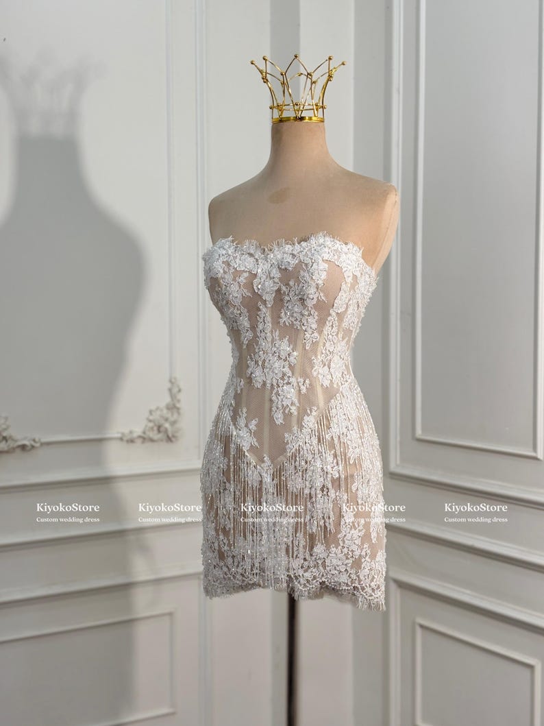May include: A strapless mini dress featuring a nude-colored base with white floral lace overlays and delicate fringe detailing along the hem. The garment is displayed on a mannequin wearing a gold-colored crown-shaped headpiece.