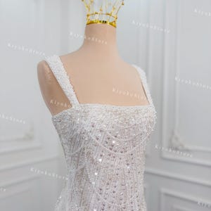May include: A white, beaded wedding dress with a square neckline and thick straps. The dress is adorned with intricate beadwork and sequins. A gold crown sits atop a mannequin head. The text "KiyokoStore" is visible.