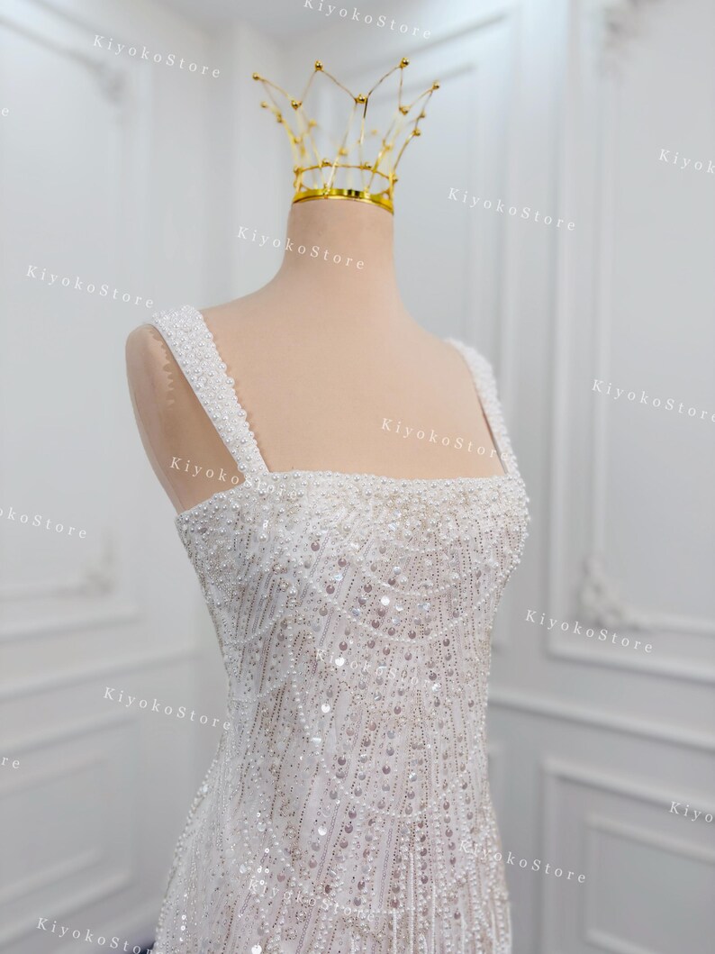 May include: A white, beaded wedding dress with a square neckline and thick straps. The dress is adorned with intricate beadwork and sequins. A gold crown sits atop a mannequin head. The text "KiyokoStore" is visible.
