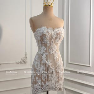 May include: A strapless mini dress featuring white floral lace appliqu&eacute;s over a nude-colored base, accented with hanging fringe details and sequins. A gold-toned wire crown sits atop the mannequin displaying the dress, which is labeled KiyokoStore.
