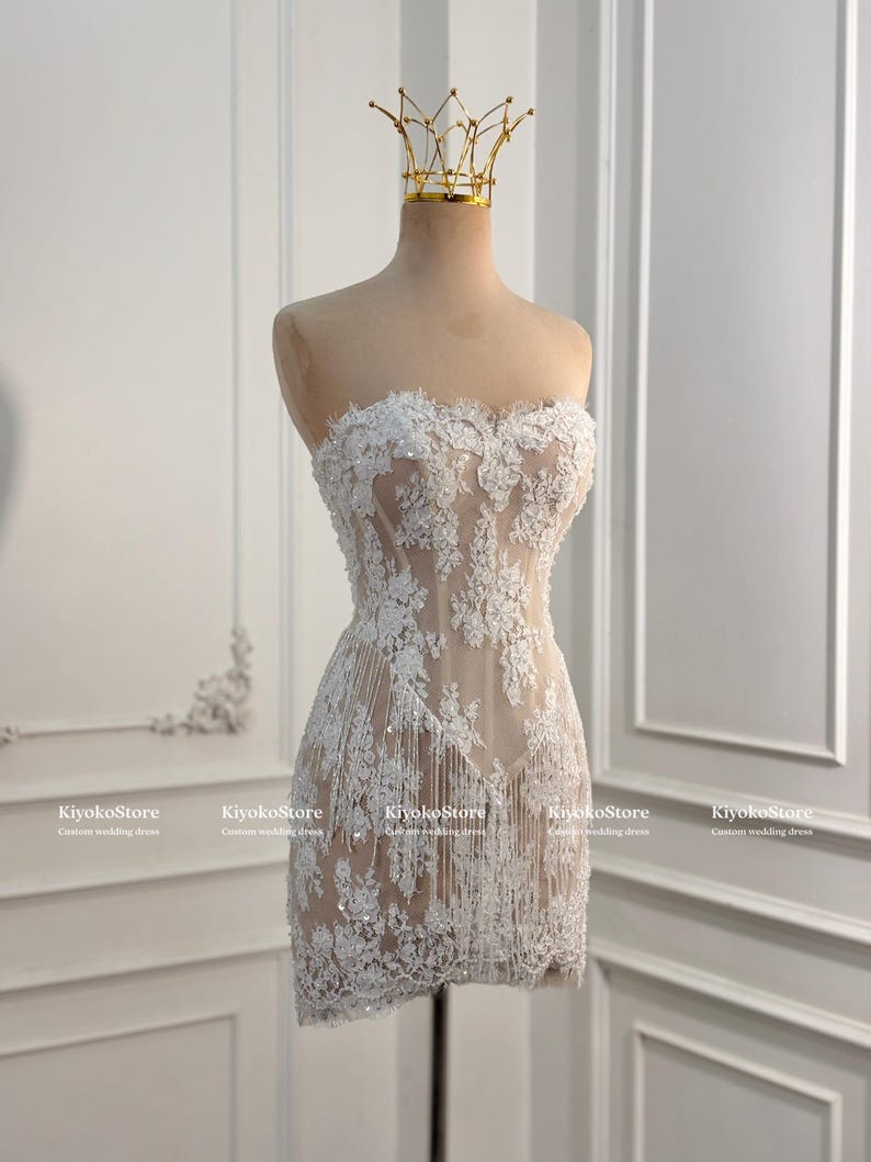 May include: A strapless mini dress featuring white floral lace appliqu&eacute;s over a nude-colored base, accented with hanging fringe details and sequins. A gold-toned wire crown sits atop the mannequin displaying the dress, which is labeled KiyokoStore.