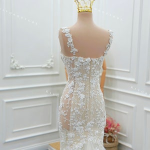 Pu&ograve; includere: Un abito da sposa senza maniche che arriva a terra, caratterizzato da una base color carne con intricati ricami in pizzo floreale bianco, un pannello laterale trasparente, un corpetto aderente, chiusura a zip e gonna svasata, sormontato da una corona in filo d'oro.