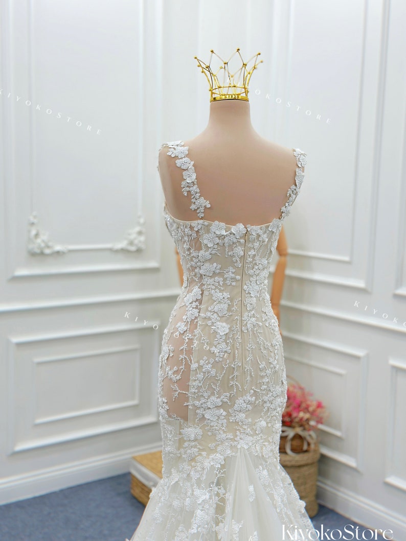 Pu&ograve; includere: Un abito da sposa senza maniche che arriva a terra, caratterizzato da una base color carne con intricati ricami in pizzo floreale bianco, un pannello laterale trasparente, un corpetto aderente, chiusura a zip e gonna svasata, sormontato da una corona in filo d'oro.
