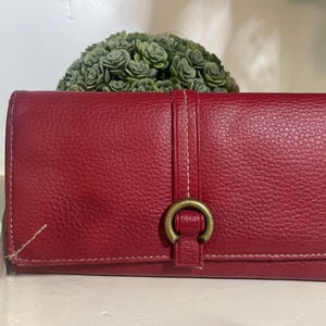 May include: A red leather wallet with a textured surface. The wallet has a vertical strap with a gold-colored ring closure. The stitching is visible in a lighter color. The wallet is rectangular and closed.