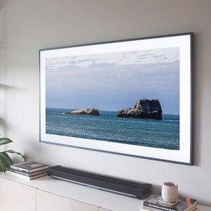 Könnte beinhalten: Ein schwarzer Fernseher mit einem Meeresbild von zwei felsigen Inseln im Ozean. Der Fernseher ist an einer Wand über einem weißen Schrank mit einer schwarzen Soundbar montiert.