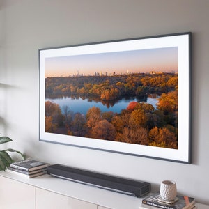 Puede incluir: Un televisor con marco negro con una imagen de un lago rodeado de árboles de otoño. Los árboles son una mezcla de naranja, amarillo y rojo. El lago refleja el cielo, que es azul claro con un toque de rosa. El horizonte de la ciudad es visible en la distancia.