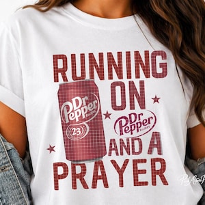May include: White t-shirt with a maroon graphic featuring a Dr. Pepper can and the text "RUNNING ON Dr Pepper AND A PRAYER". The Dr. Pepper can is red with white text and the number 23.