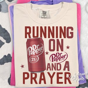 May include: Cream-colored t-shirt with the text "RUNNING ON Dr Pepper AND A PRAYER" in maroon. A maroon Dr Pepper can graphic is on the shirt. The shirt is folded on top of pink and purple shirts.