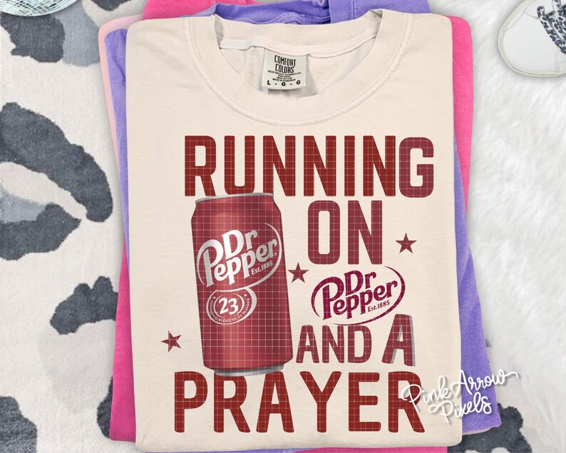 May include: Cream-colored t-shirt with the text "RUNNING ON Dr Pepper AND A PRAYER" in maroon. A maroon Dr Pepper can graphic is on the shirt. The shirt is folded on top of pink and purple shirts.