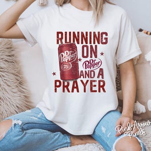 May include: Cream-colored t-shirt featuring a distressed graphic of a Dr. Pepper can and the words "RUNNING ON Dr. Pepper AND A PRAYER" in red. The shirt has short sleeves and a relaxed fit.