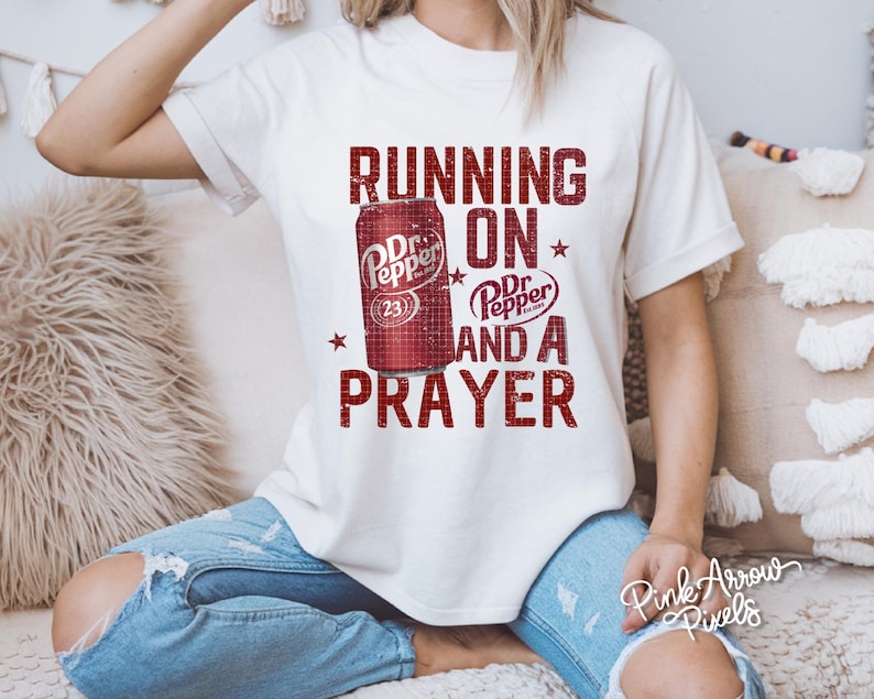 May include: Cream-colored t-shirt featuring a distressed graphic of a Dr. Pepper can and the words "RUNNING ON Dr. Pepper AND A PRAYER" in red. The shirt has short sleeves and a relaxed fit.