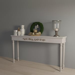 Könnte beinhalten: Ein weißer Konsolentisch mit dekorativen Beinen und einer detaillierten Frontplatte. Der Tisch ist mit einem grünen Kranz, goldenen Ornamenten, einem Glaskerzenhalter und weißen dekorativen Behältern geschmückt. Der Tisch steht vor einer grauen Wand.