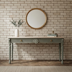 May include: A sage green console table with a light wood top, ornate detailing, and brass hardware. A round gold mirror hangs above the table against a brick wall. A white vase with greenery and books are on the table.