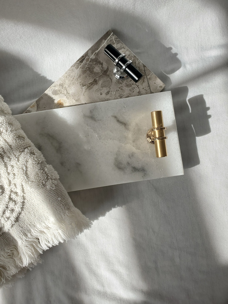 May include: Two marble serving trays with metal handles. One is rectangular with a white and gray pattern and gold handles. The other is triangular with a gray and brown pattern and black handles. A white towel is in the corner.