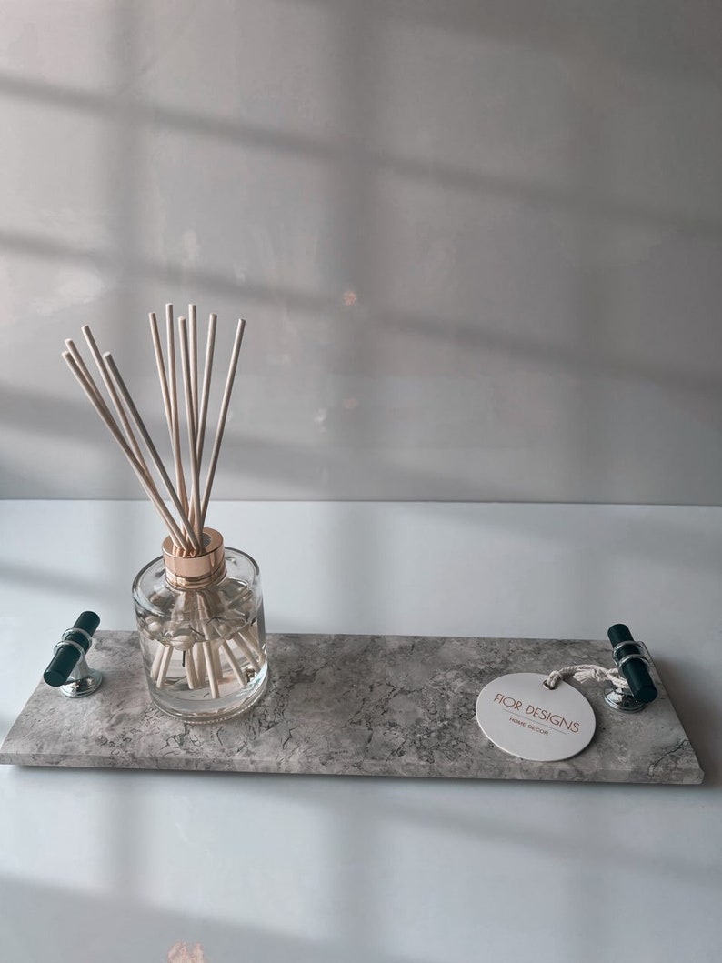 May include: A rectangular gray marble tray with a clear glass diffuser bottle and wooden reeds. The tray has two black and silver handles and a white tag that reads "FIOR DESIGNS HOME DECOR".