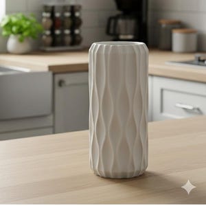 May include: A tall, off-white ceramic vase with a textured, wave-like pattern. The cylindrical vase is displayed on a light-colored wooden surface. The kitchen setting includes a coffee maker and spice rack in the background.