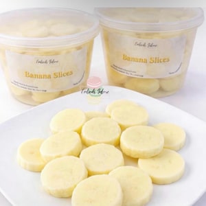 Realistic banana slice wax melts on a white plate with additional pieces for fuller appearance, pale yellow smooth molded wax embeds, Embeds Galore banana slices in clear containers in background, product size reference with coin, dessert wax melts for candles and wax melt crafting