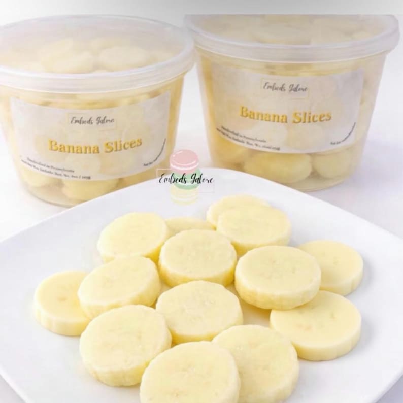 Realistic banana slice wax melts on a white plate with additional pieces for fuller appearance, pale yellow smooth molded wax embeds, Embeds Galore banana slices in clear containers in background, product size reference with coin, dessert wax melts for candles and wax melt crafting