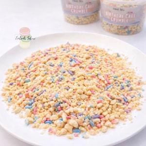 White plate filled with birthday cake–style wax crumble embeds featuring pastel sprinkles, with Embeds Galore “Birthday Cake Crumble” containers blurred in the background on a white surface.