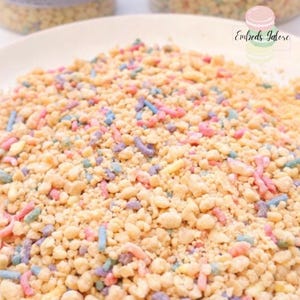Close-up of birthday cake wax crumble embeds with pastel sprinkle pieces spread across a white plate, branded Embeds Galore, photographed on a clean white background.