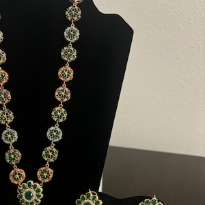 May include: A gold necklace and earrings set featuring green, red, blue, and white stones. The necklace has a large pendant with a floral design. The earrings are also floral and have pearl drops.