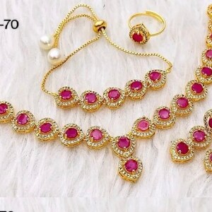 May include: A gold-tone jewelry set with pink gemstones. The set includes a necklace, bracelet, earrings, and a ring. The necklace and bracelet feature a teardrop design with a large pink gemstone in the center. The earrings are long and dangle, with three pink gemstones each. The ring has a single pink gemstone.