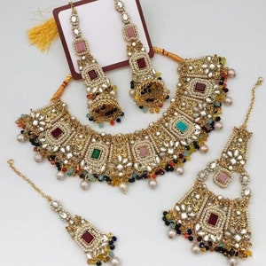 May include: A gold-tone bridal jewellery set with a multi-strand necklace, earrings, and a tikka. The necklace features a large, ornate pendant with colourful stones and mirrors. The earrings are long and dangle, with a similar design to the necklace. The tikka is a small, ornate piece that sits on the forehead.