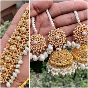 May include: A set of gold-tone earrings with white pearl accents. The earrings feature a large round pendant with a smaller round pendant hanging from it. The earrings are strung on a white pearl chain.