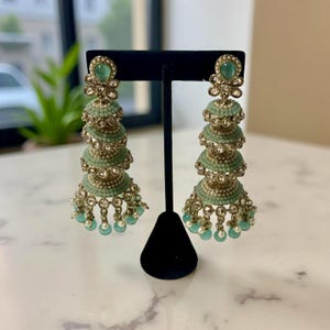 May include: Mint green and gold chandelier earrings. The earrings feature a tiered design with small, round embellishments and dangling beads. The earrings are displayed on a black stand, showcasing their intricate details and elegant style.