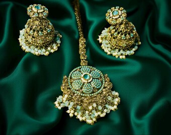 Puede incluir: Un conjunto de joyas dorado y ornamentado, que incluye una pieza para la cabeza y pendientes a juego. El conjunto está adornado con piedras de color turquesa y pequeñas cuentas blancas, sobre un fondo de satén verde esmeralda.