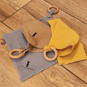 May include: Three muslin baby bibs with wooden teething rings. The bibs are in blue, yellow, and brown with the text "Schmuddelkind" printed on them.