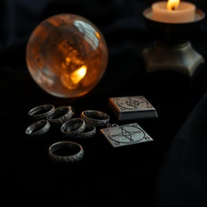 May include: A collection of silver rings, pendants, and a small box with intricate designs, arranged on a black surface. A lit candle in a holder and a glowing crystal ball are in the background, creating a mystical ambiance.