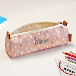 May include: Pink pencil case with a white raindrop pattern and the name "Marie" embroidered in gray. The case is open and lying on a white surface.