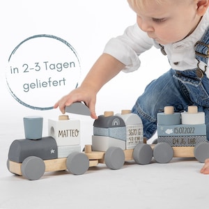 Könnte beinhalten: Ein grauer und weißer Holzeisenbahnzug mit einem Stern auf der Lokomotive. Der Zug hat fünf Waggons, jeder mit einem anderen Design. Der Waggon, der der Lokomotive am nächsten ist, trägt den Namen "MATTEO". Der nächste Waggon hat einen Regenbogen und den Text "3670 G 53 CM". Der dritte Waggon hat eine Wolke und den Text "15.07.2022". Der vierte Waggon hat ein Zickzackmuster und den Text "04:18 Uhr". Der fünfte Waggon ist ein schlichter grauer Block. Ein Kind spielt mit dem Zug.