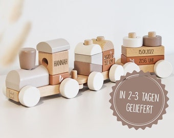 May include: A wooden toy train with a gray and brown color scheme. The train has five cars, each with a different design. The first car has the name "HANNAH" on it. The second car has the text "3670 G 53 CM" on it. The third car has the date "15.01.2022" and the time "21:56 UHR" on it. The train is on a white background. The text "IN 2-3 TAGEN GELIEFERT" is in a circle on the bottom right of the image.