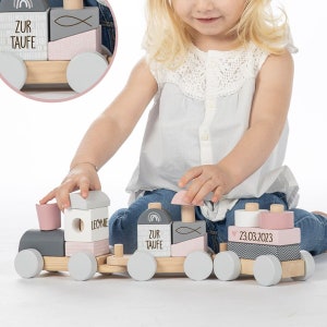 Könnte beinhalten: Ein Holzspielzeugzug mit grauen, weißen und rosa Blöcken. Die Blöcke haben Text und dekorative Elemente. Der Zug hat graue Räder und ist für Kinder konzipiert. Der Text auf den Blöcken enthält die Wörter "ZUR TAUFE" und den Namen "LEONIE".