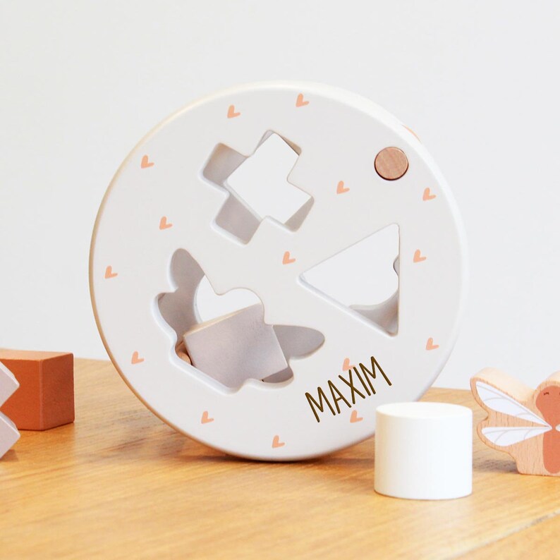 May include: White wooden shape sorter toy with a personalized name "MAXIM" and pink hearts. The toy has different shaped cutouts for the child to put the blocks through.