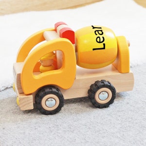Può includere: Un camioncino di cemento giocattolo in legno giallo con un tamburo rotante. Il tamburo è etichettato "Lear". Il camioncino ha ruote nere e una piccola figura di una persona nella cabina.