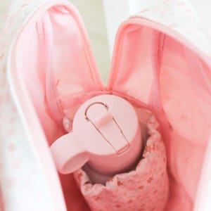May include: A light pink insulated water bottle with a handle and a flip-top lid is nestled inside a matching pink bag. The bag has a floral pattern and a zippered opening. The interior of the bag is also pink.