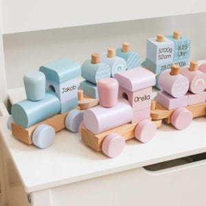 Könnte beinhalten: Holzspielzeug-Zugset in Pastellfarben. Die Waggons sind hellblau, rosa und lavendelfarben, mit Holzrädern und Akzenten. Einige Waggons tragen Namen, einer Geburtsdaten. Ein bezauberndes und lehrreiches Spielzeug.